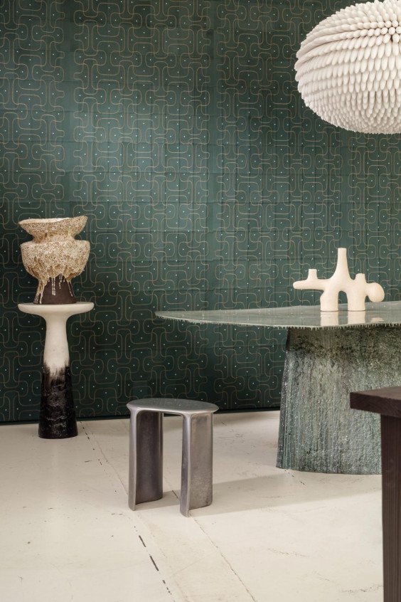 Papier peint géométrique vert foncé Corium Jaseran dans une salle à manger élégante avec table en verre et objets sculpturaux.